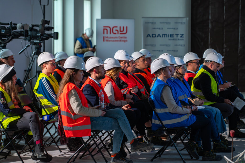 Goście podczas konferencji prasowej MGW, ujęcie od boku. Goście siedzą na składanych krzesełkach i posiadają kamizelki odblaskowe oraz kaski ochronne. W tle baner MGW i Adamietz.
